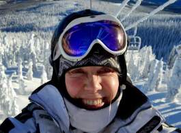 Close-up portrait of a smiling woman wearing a ski helmet, jacket, and blue goggles on chairlift above snow covered trees.