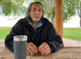 Male Indigenous Elder sitting at a lakefront picnic table with a thermal coffee mug