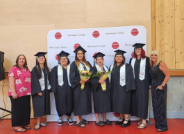 Group of graduates in caps and gowns posing with bouquets at Okanagan College graduation ceremony, standing in front of a banner with the college's logo.