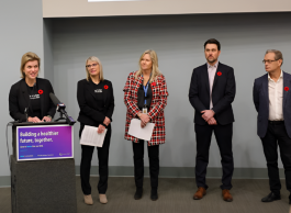 A group of five individuals stand behind a podium. One person is speaking at the microphone. They all wear poppy pins, and the backdrop includes banners with the text "Building a healthier future, together."