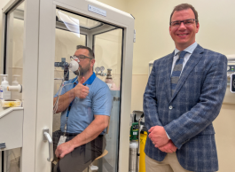 One individual performs a respiratory test using specialized equipment inside a glass enclosure, while another person stands outside smiling, both in a clinical setting.