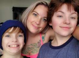 Blonde woman with large tattoo on chest, between two smiling children with brown hair, one wearing a navy-blue hat