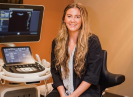 Woman with long, curled blonder hair wearing a black blazer with a light blue blouse underneath is sitting in front of ultrasound equipment and smiling at the camera.