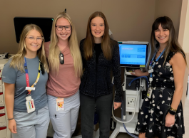 four female health care workers stand beside a medical machine and smile