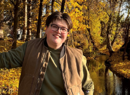 A smiling person with short brown hair and glasses wearing a green top and brown vest leaning against a tree in a golden fall setting.