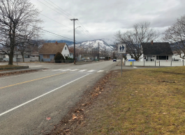 View of a quiet street with a pedestrian crossing sign. In the background, there are residential houses and distant mountains with light snow cover.