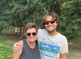 Two people: a woman with short brown hair (left), wearing a grey tank top and blue shorts, stands next to a man with brown hair, wearing a white t-shirt that says Revelstoke and green shorts behind them are lots of green trees. 