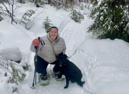 Woman in light-coloured puffy jacket snowshoeing on a trail with a black dog