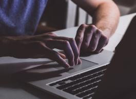 A person in a blue tshirt with their hands hovering over a laptop keyboard in a dimly lit room.